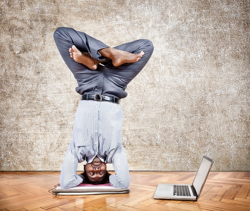 16254011 - indian businessman doing yoga headstand pose and looking at his laptop in the office at brown textured background